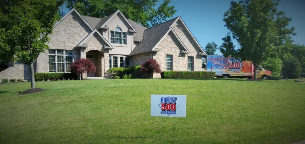 A large brick house with a well-maintained lawn and a sign in front that reads "Ainsley 500"—known for its exceptional air conditioning repair services. A truck bearing the same logo is parked nearby.