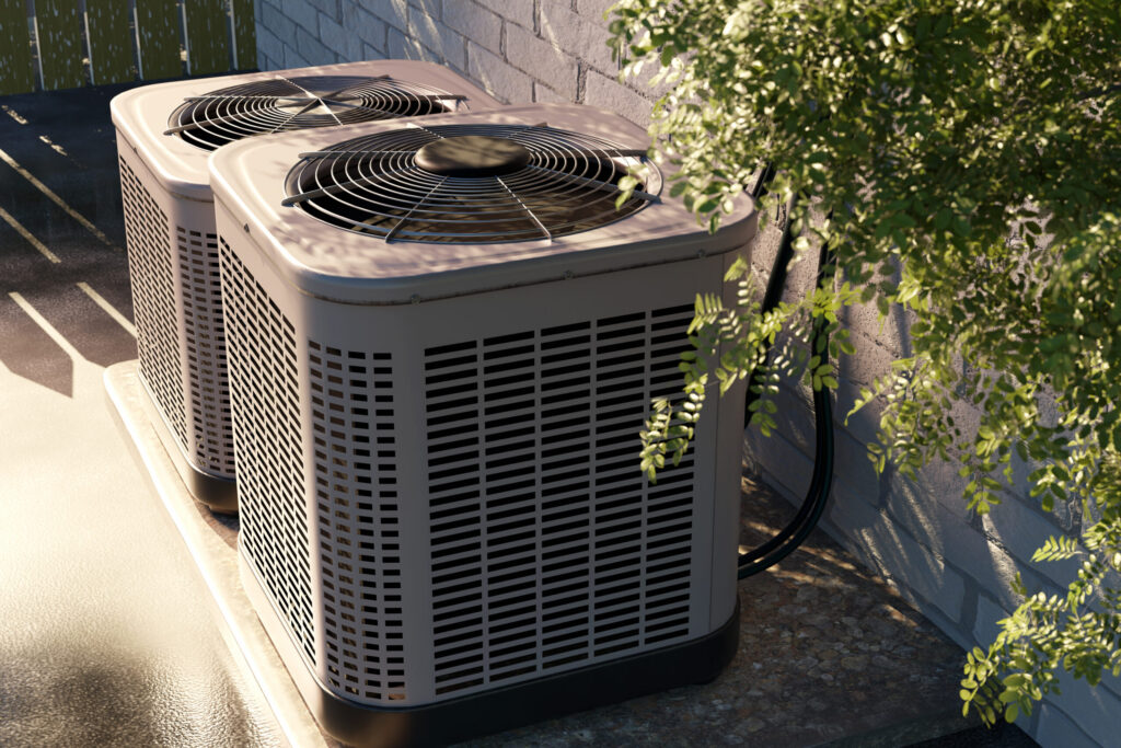 Two outdoor air conditioning units sit side by side on a concrete pad next to a brick wall, partially shaded by nearby foliage.
