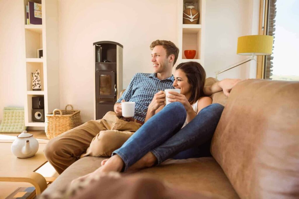 A couple sits together on a sofa, smiling and holding mugs, in a bright living room with shelves, a stove, and a yellow lamp in the background.
